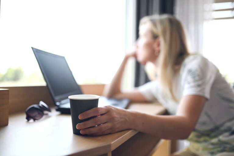 sad unhappy blonde woman holding a paper cup of coffee and sitting and dreaming at the working desk